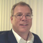 A man with short light brown hair and glasses, wearing a dark suit jacket and white shirt, smiling while sitting indoors in front of vertical blinds.