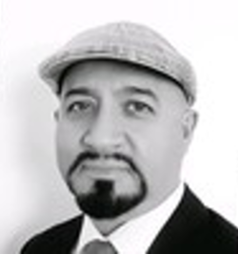 Black and white portrait of a man wearing a flat cap, suit, and tie, with a trimmed beard and mustache, looking directly at the camera against a plain background.