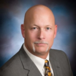 A middle-aged bald man with a mustache wearing a gray suit, white shirt, and patterned tie poses for a professional portrait against a blue and purple gradient background.