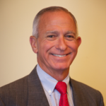 A middle-aged man with short gray hair smiles, wearing a gray suit, blue checked shirt, and red polka-dot tie, standing in front of a plain light-colored background.