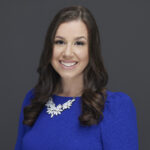 A woman with long brown hair, wearing a bright blue dress and a white floral necklace, smiles at the camera against a plain dark gray background.
