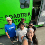 Five smiling people pose for a group selfie outside a bright green van with “ROADTRIP NATION” written on the side. The group stands on pavement near the open side door of the van.