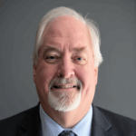 A smiling older man with white hair and a white goatee wears a dark plaid suit, light blue shirt, and patterned tie against a plain gray background.
